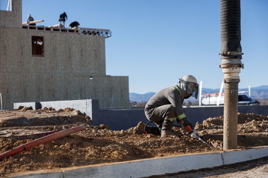 Worker on a bare dirt pad beside a poured concrete stem wall, with a framed house behind